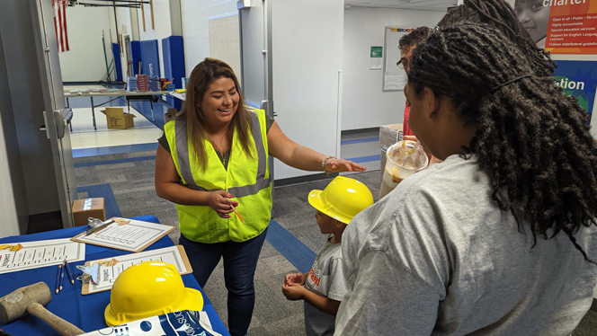 Children wearing hard hats and talking to a woman in a safety vest.
