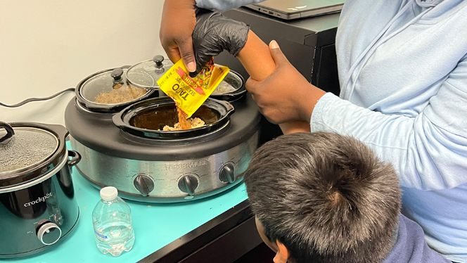 An adult helping a student add taco seasoning to one of the crockpots.